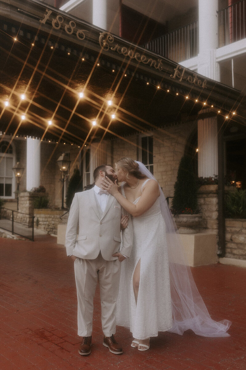 A bride and groom in wedding attire stand in front of the Crescent Hotel in Eureka Springs, Arkansas. They are kissing in front of the awning that says "1886 Crescent Hotel" in ornate gold lettering. The lights underneath the awning create a magical starburst effect.