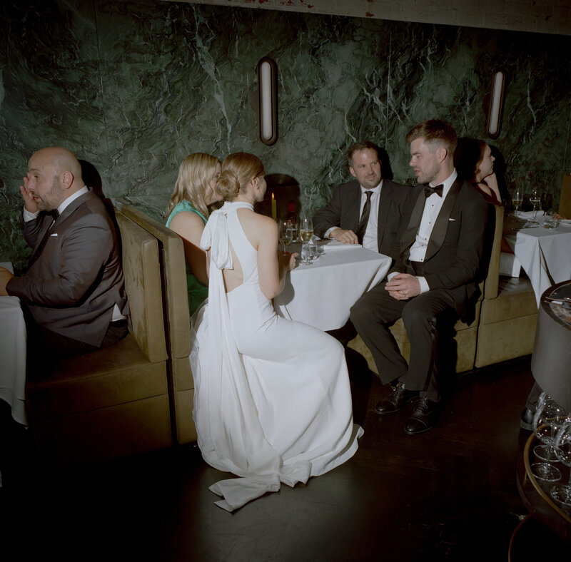 A sophisticated wedding scene at Cutler and Co in Fitzroy, featuring a stylish group of guests seated in an intimate booth. The bride, dressed in an elegant white gown with a flowing train and halter-neck design, sits gracefully with her back to the camera. Her hair is swept up into a neat bun, emphasizing the minimalist and modern design of her dress. The groom, seated across from her in a classic black tuxedo with a bow tie, is engaged in relaxed conversation. Other guests, also dressed in formal black-tie attire, occupy the booth, contributing to the chic, upscale atmosphere of the reception. The booth seating, upholstered in luxurious olive-green velvet, is set against a striking emerald-green marble wall, which adds a touch of opulence to the venue's decor. On the tables are wine glasses, half-filled, and small candles, casting soft, romantic lighting across the scene. The dark hardwood floors and ambient lighting enhance the intimate and elegant vibe of this Melbourne wedding reception at Cutler and Co.