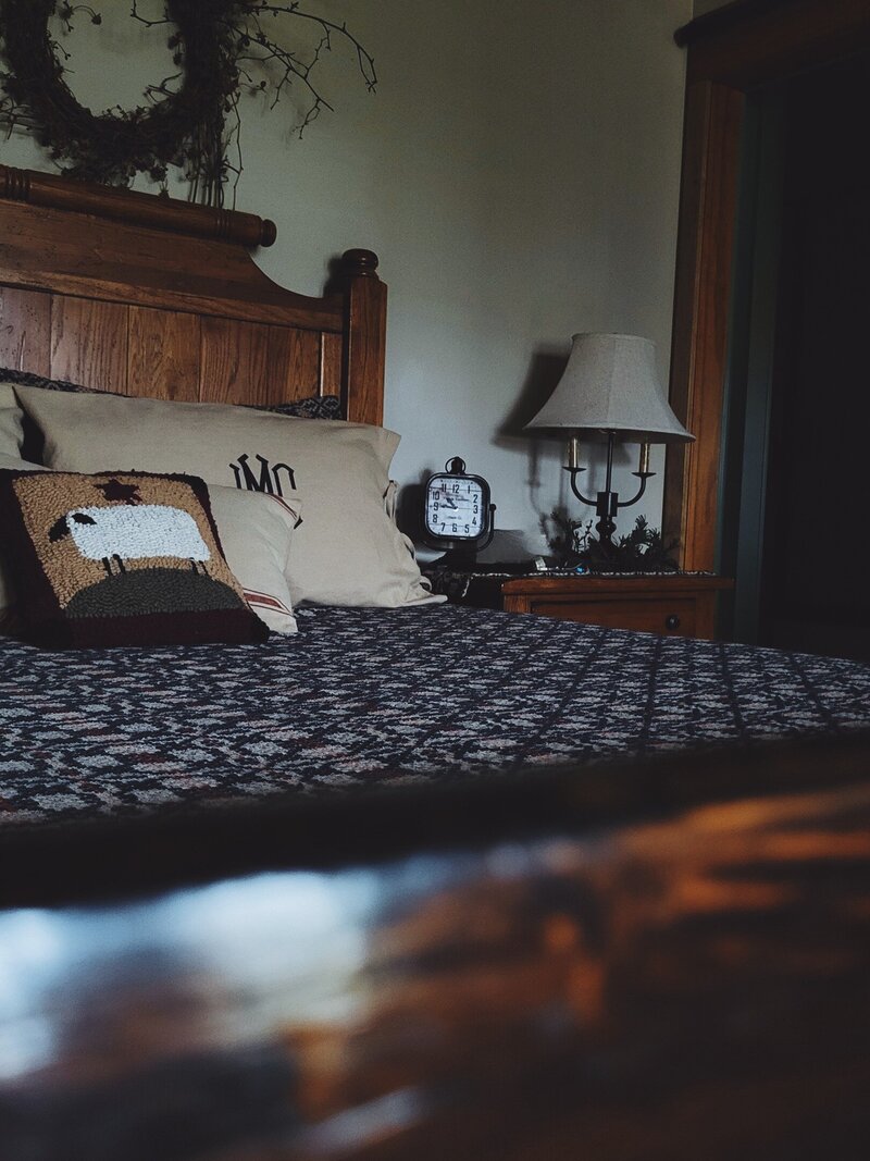 Tall oak bed, with early American textile & monogrammed pillow covers and a matching nightstand with oval cream candleabra lamp