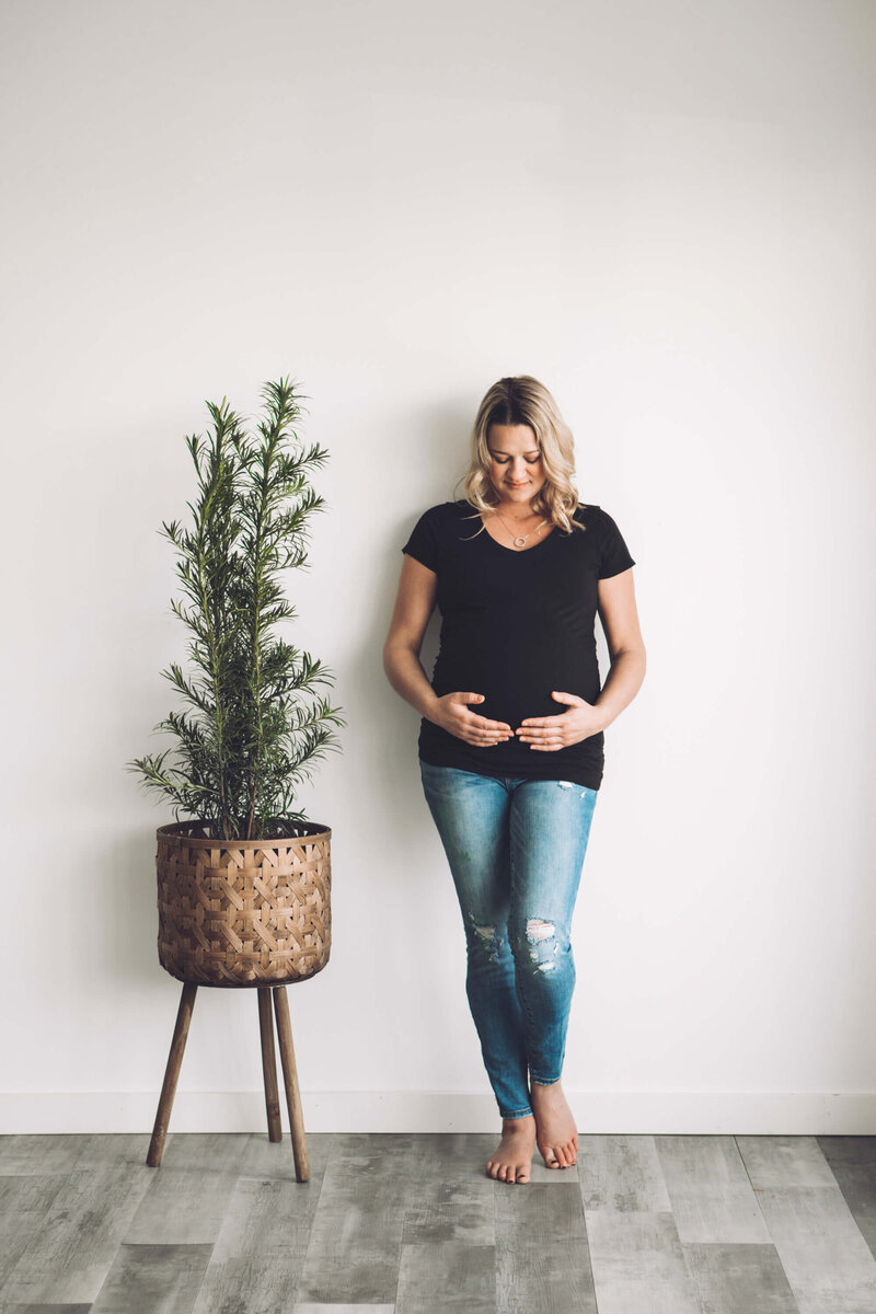 expectant mom in jeans leans on wall by a plant