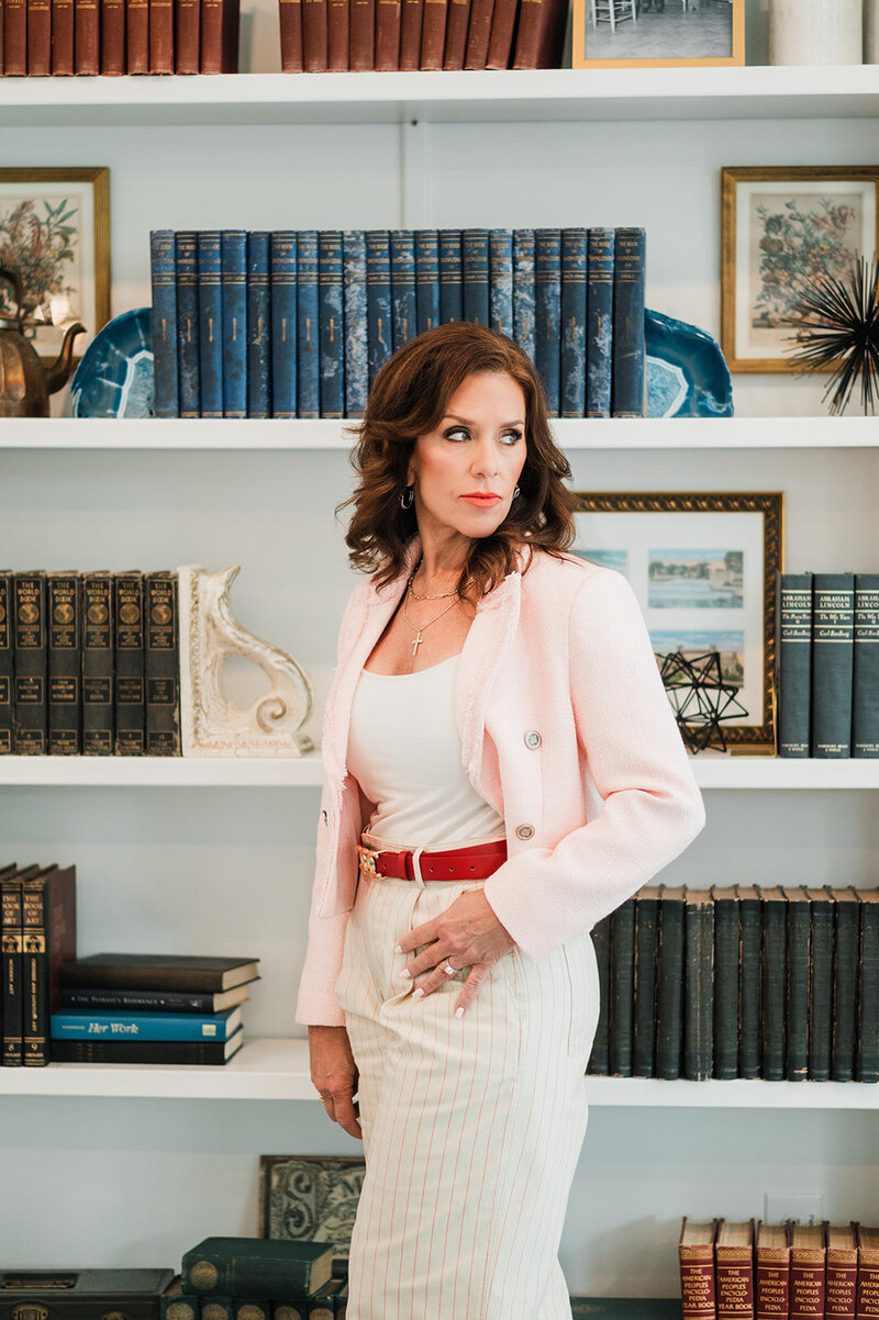 A wedding planner stands poised in front of a bookshelf, exuding elegance and professionalism, embodying the sophisticated style of Socially Significant Events.