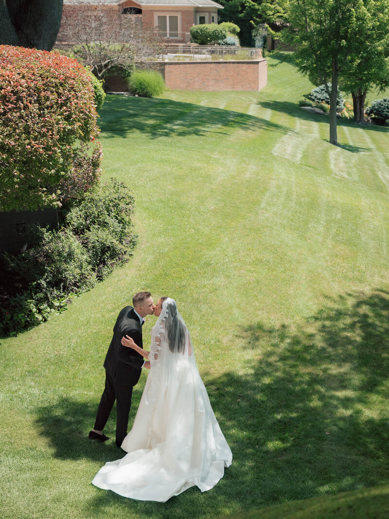 Bride and groom kiss on lush green lawn in full sunlight after outdoor ceremony