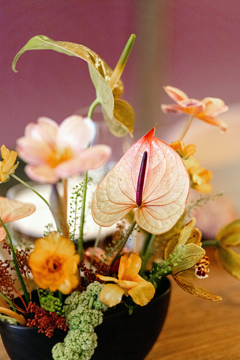 Vibrant floral arrangement featuring anthuriums, orchids, and textured greenery in a black vase with a warm-toned background.