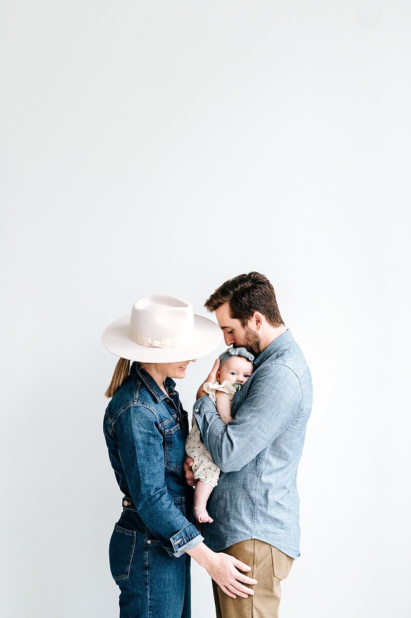 mom and dad snuggle baby girl in front of a white wall