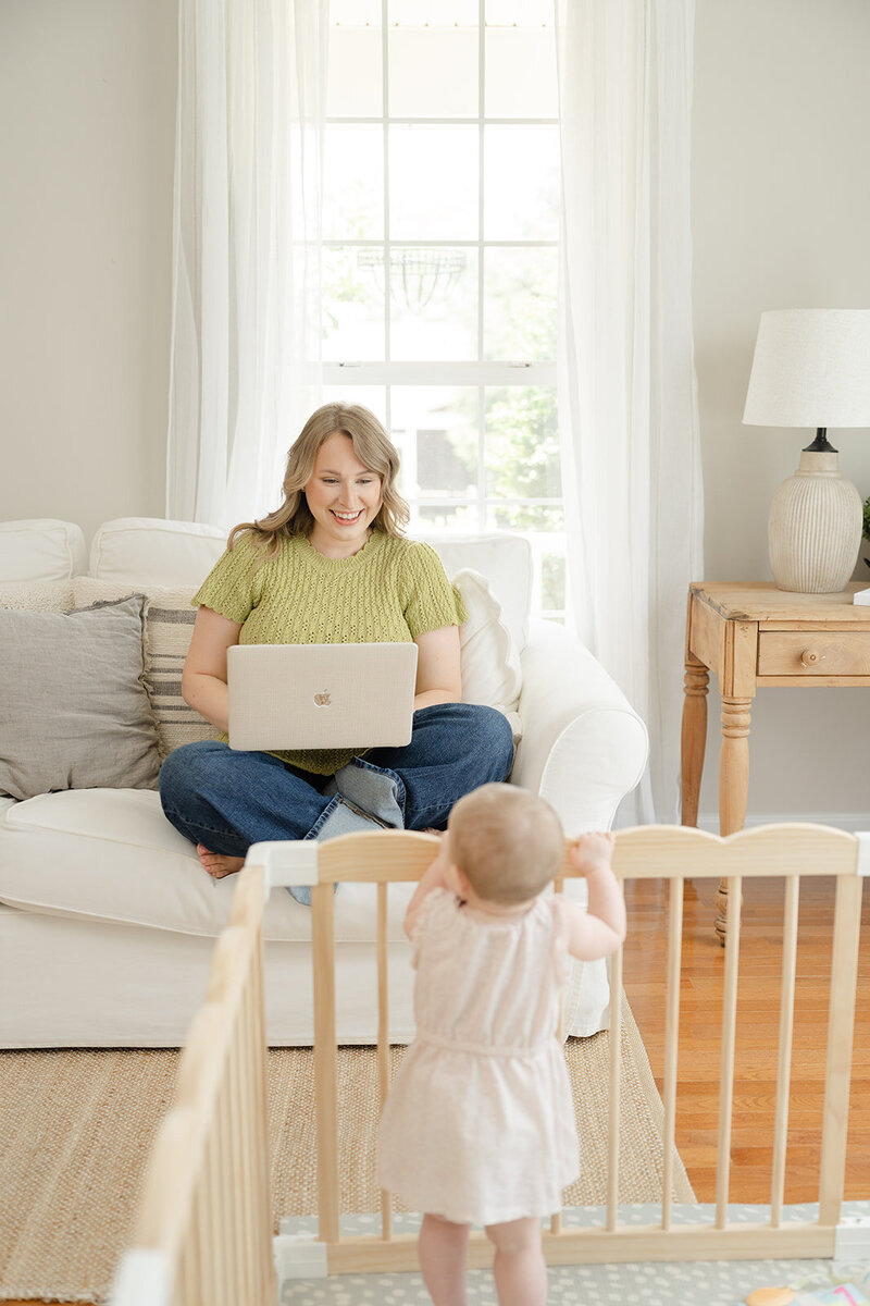 Mother working on her Macbook Pro on a white couch sitting criss-cross while looking down at baby girl playing in wooden play area