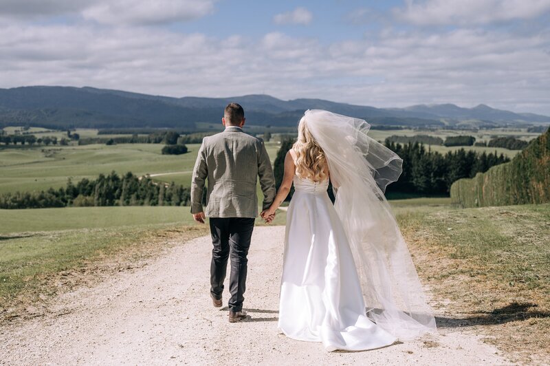 a bride and groom walk away down gravel road overlooking green fields of waimumu near gore in new zealand