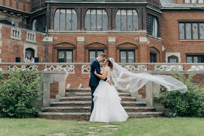 Bride and groom smile at each  other while the bride's veil blows in the wind at Rollins Mansion . Photo  by Anna brace a wedding photographer in iowa.