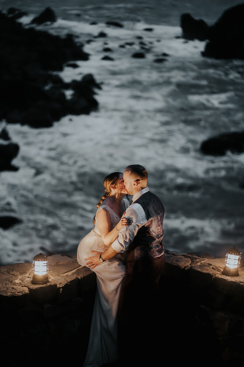 a couple in wedding attire embrace as the bride sits on a stone wall. they are illuminated by the warm glow of lanterns as ty kiss. Taken from a higher vantage point by their adventure elopement photographers, we see the crashing, ocean waves swirling beneath them.