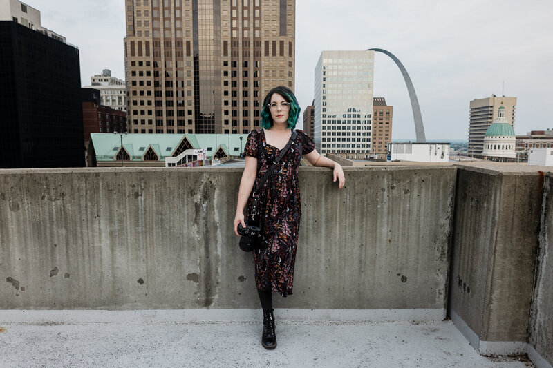 Heather Bennett, owner of Bee Brook Photography, leans against a concrete wall with her camera in her right hand on the rooftop of the Kiener West Parking Garage in St. Louis. The St. Louis Gateway Arch and skyscrapers can be seen in the background.