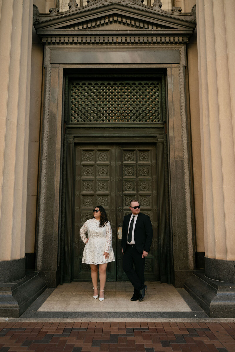 A stylish couple in sunglasses stands in front of an ornate, large wooden door framed by tall columns. The mood is elegant and sophisticated.