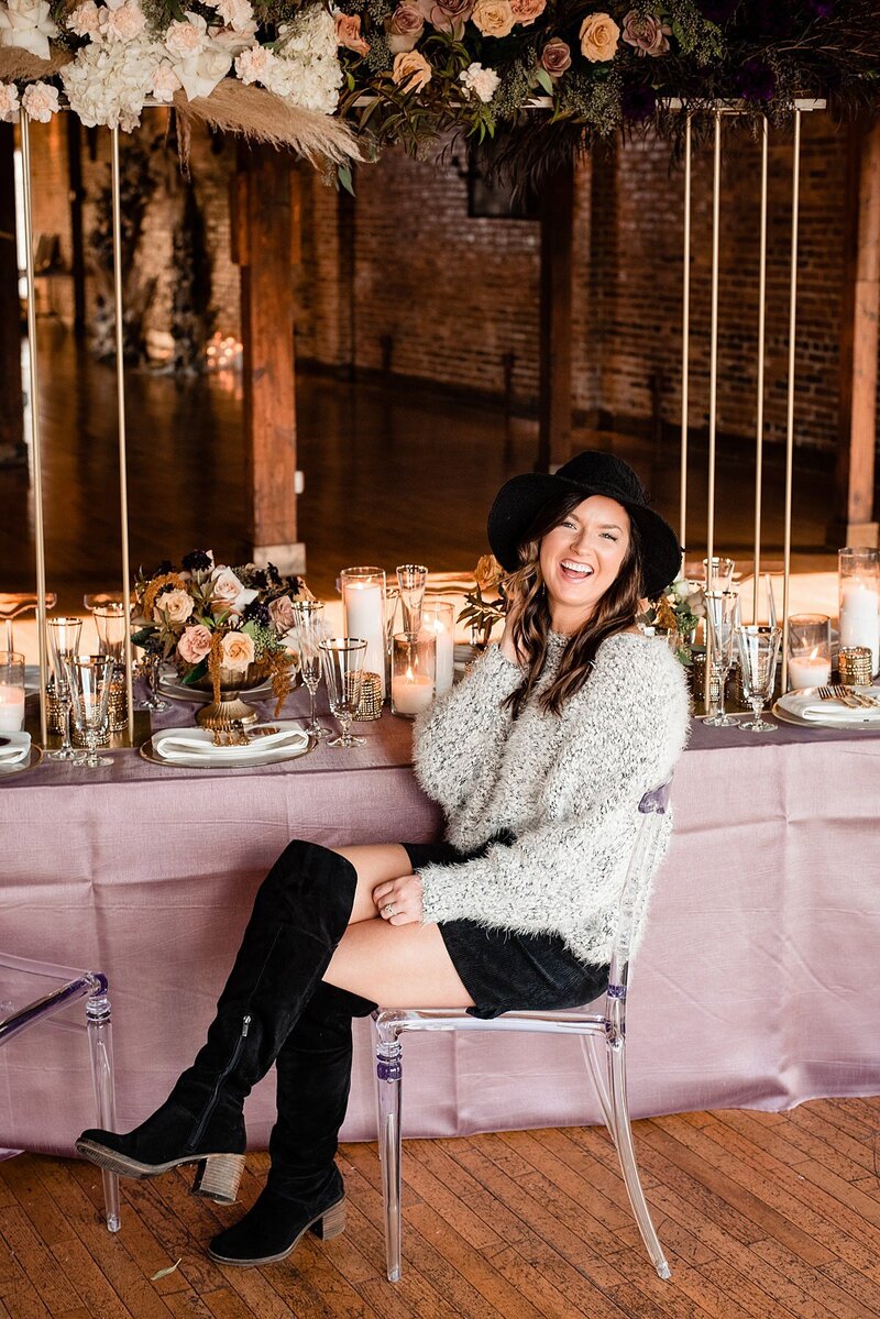 Wedding planner and florist wearing a cute fall outfit and black floppy hat sitting down and laughing toward camera with wedding tablescape at her back