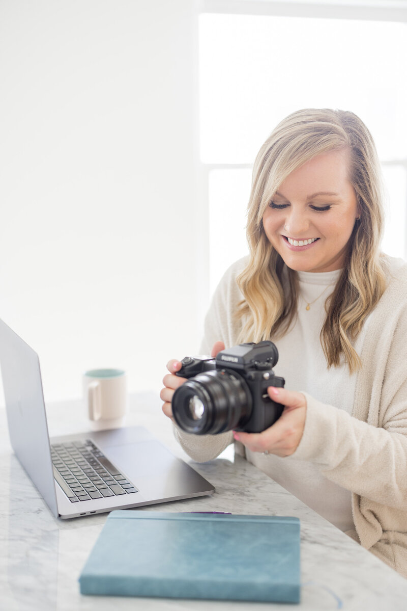 Krista Marie, a brand photographer for women, picking up her camera and smiling while working on her laptop