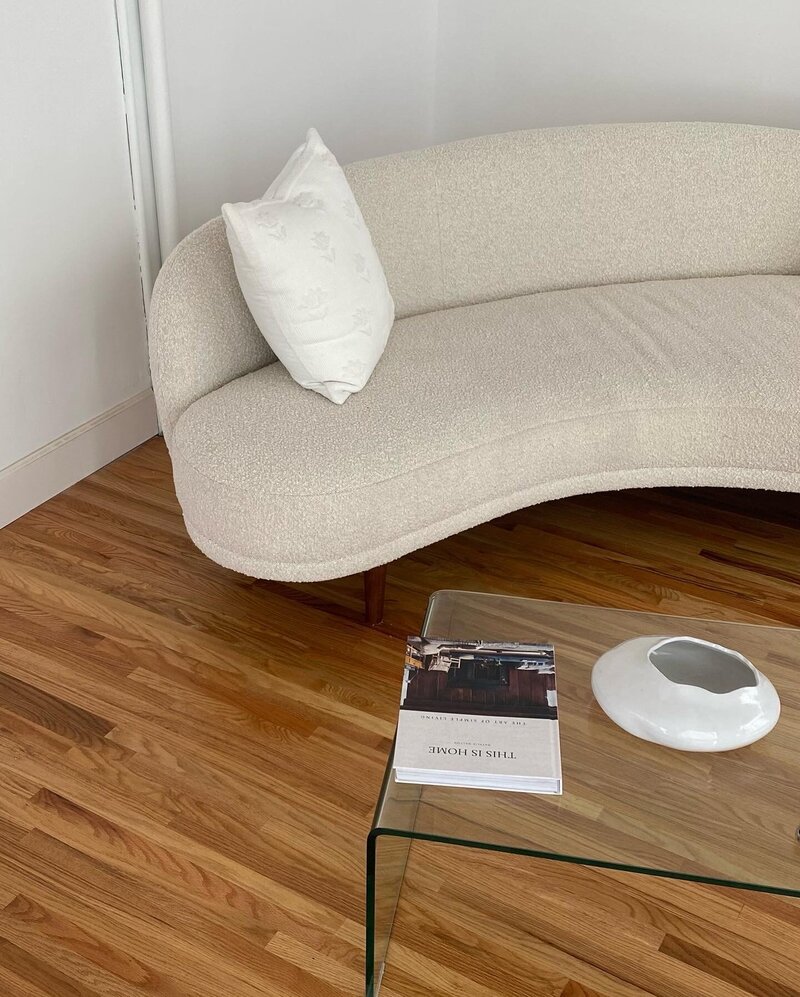 Curved beige sofa with a white pillow in a minimalist room. Glass coffee table holds a white ceramic piece and a book. Warm, cozy ambiance.