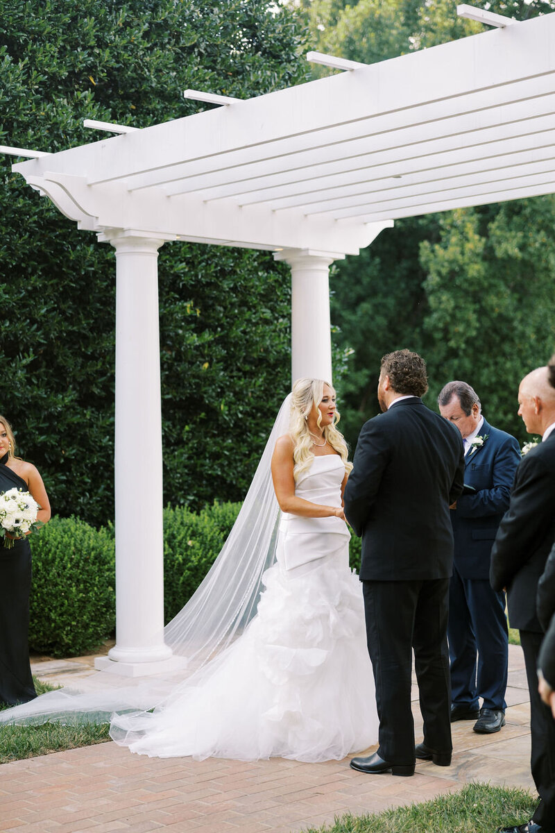 Kailea and Zach holding hands under an archway on the lawn of Boxwood Estate. During their ceremony, Kailea is wearing a white gown with white veil while Zach is wearing a classic suit. 