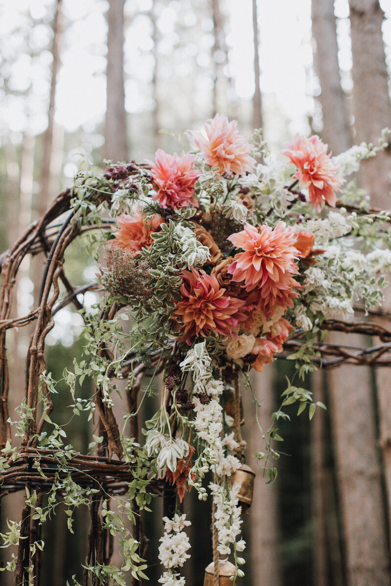 A detail of a rustic wedding ceremony arch with peach dahlias celosia roses and larkspur standing in a pine forest in upstate New York