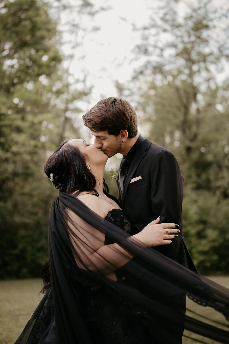 bride with black veil kissing groom in forrest