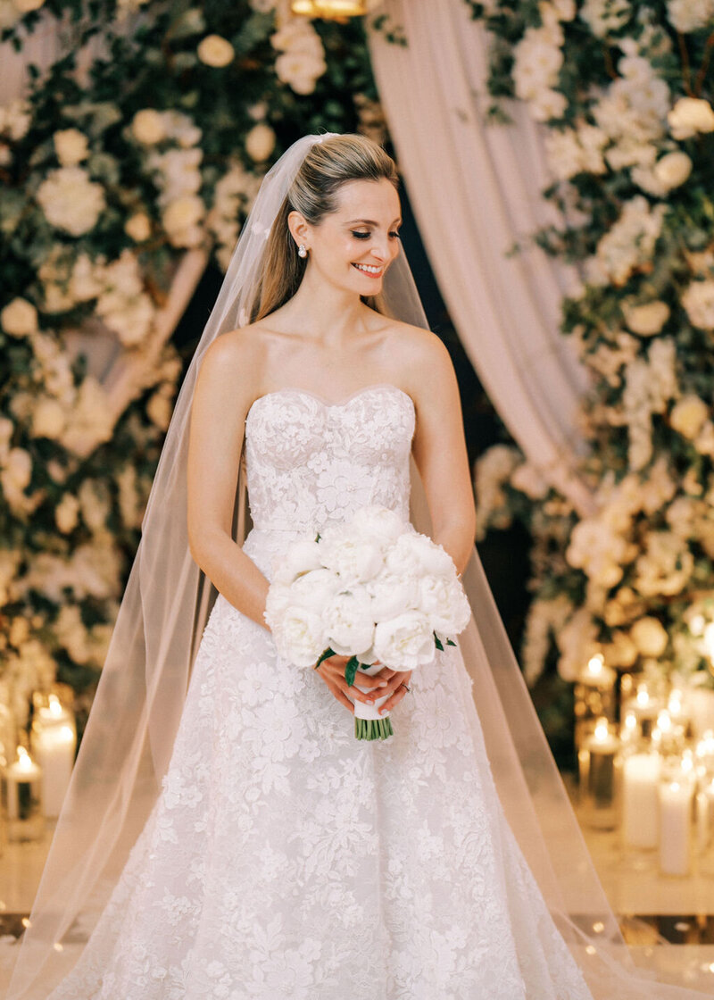 Bride in an elegant strapless wedding gown, holding a bouquet of white flowers, poses in a luxuriously decorated ballroom with golden drapes and a lavish floral arrangement in the background at The Langham Chicago.