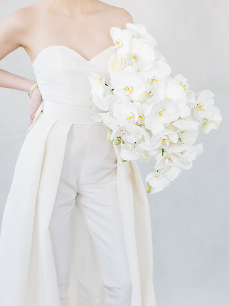 bride wearing pant suit with cape wedding dress holding an all white orchid bouquet