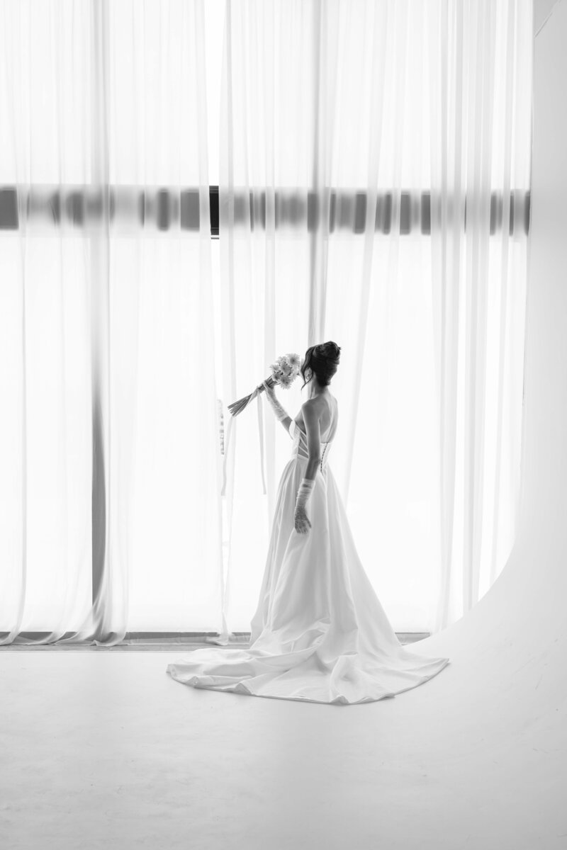 black and white photo of a bride smelling her wedding boquete looking through a brigth window