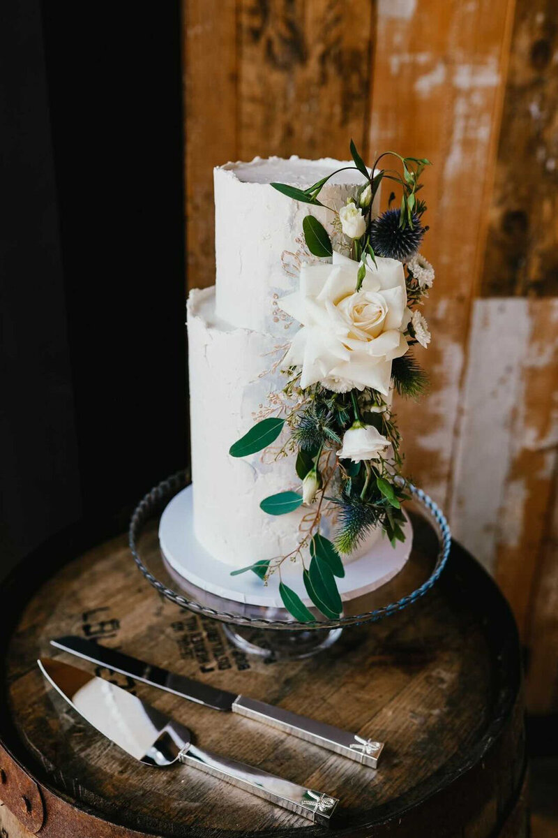 Two tier wedding cake in a rustic style with textured buttercream and  fresh florals - white roses and loose trailing foliage with splashes of gold sitting on a wooden barrel