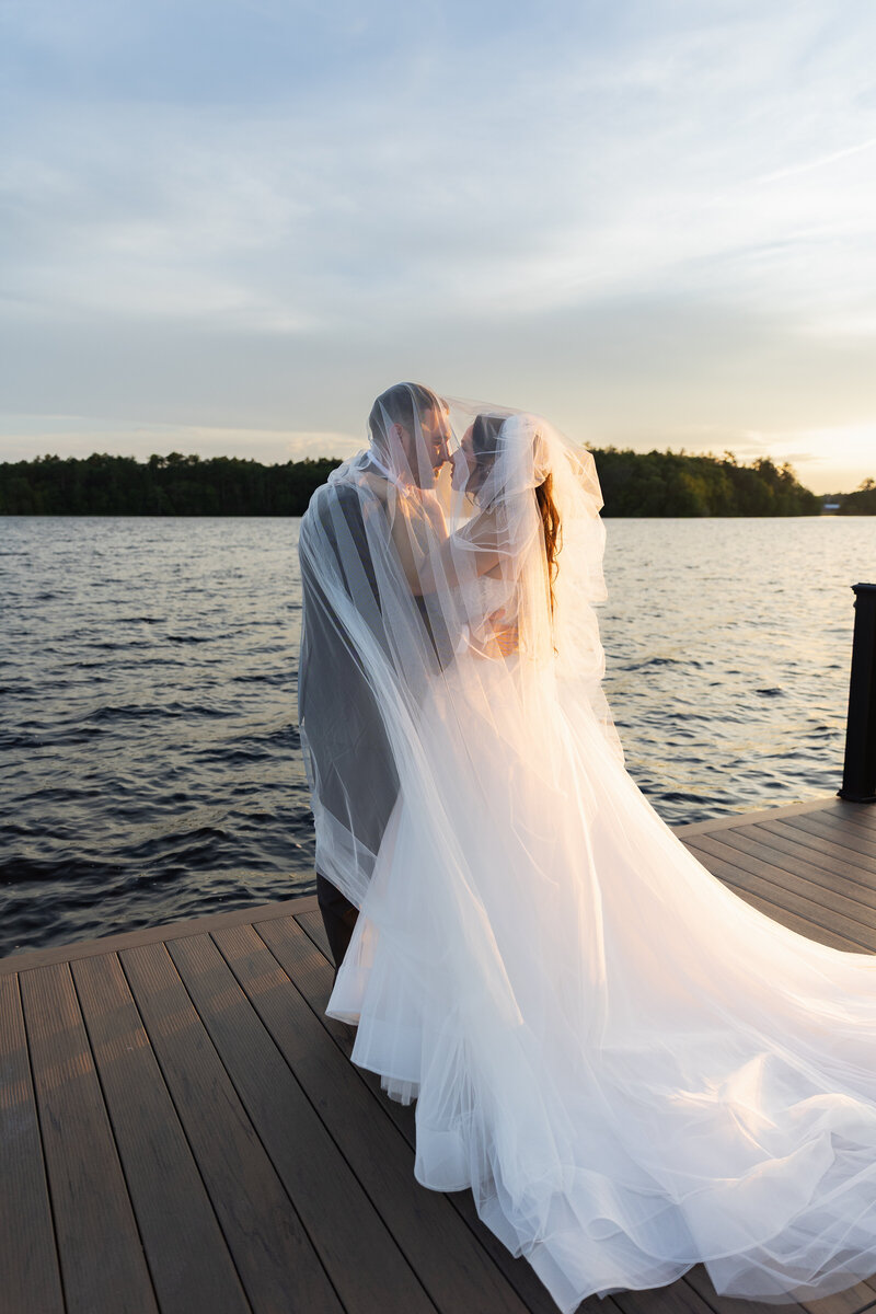 The Lakehouse Inn MA  | Kelsey Sheehan Photography Timeless Rhode Island Weddings | Bride and groom embrace on a lakeside pier at sunset, veils softly draped around them. The serene water and gentle light create a romantic atmosphere.