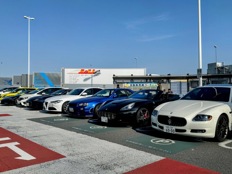 A row of high-end sports cars including Ferraris, Maseratis, and Japanese imports parked at Tatsumi Parking Area in Tokyo under a clear blue sky.