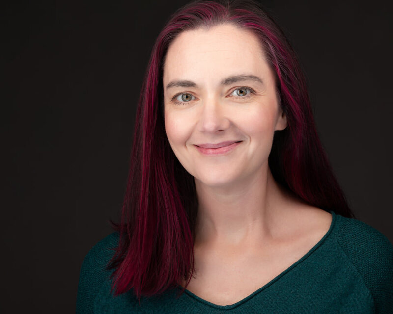 Fort Mill SC boy mom portrait photographer Alicia Insley Smiht smiles in her portrait wearing a green sweater posing in front of a black background in a studio setting with pink hair