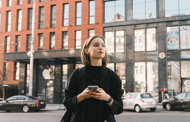 A young woman in a black coat and pants stands in front of a modern building with large glass windows, holding a smartphone in both hands and looking to the side.