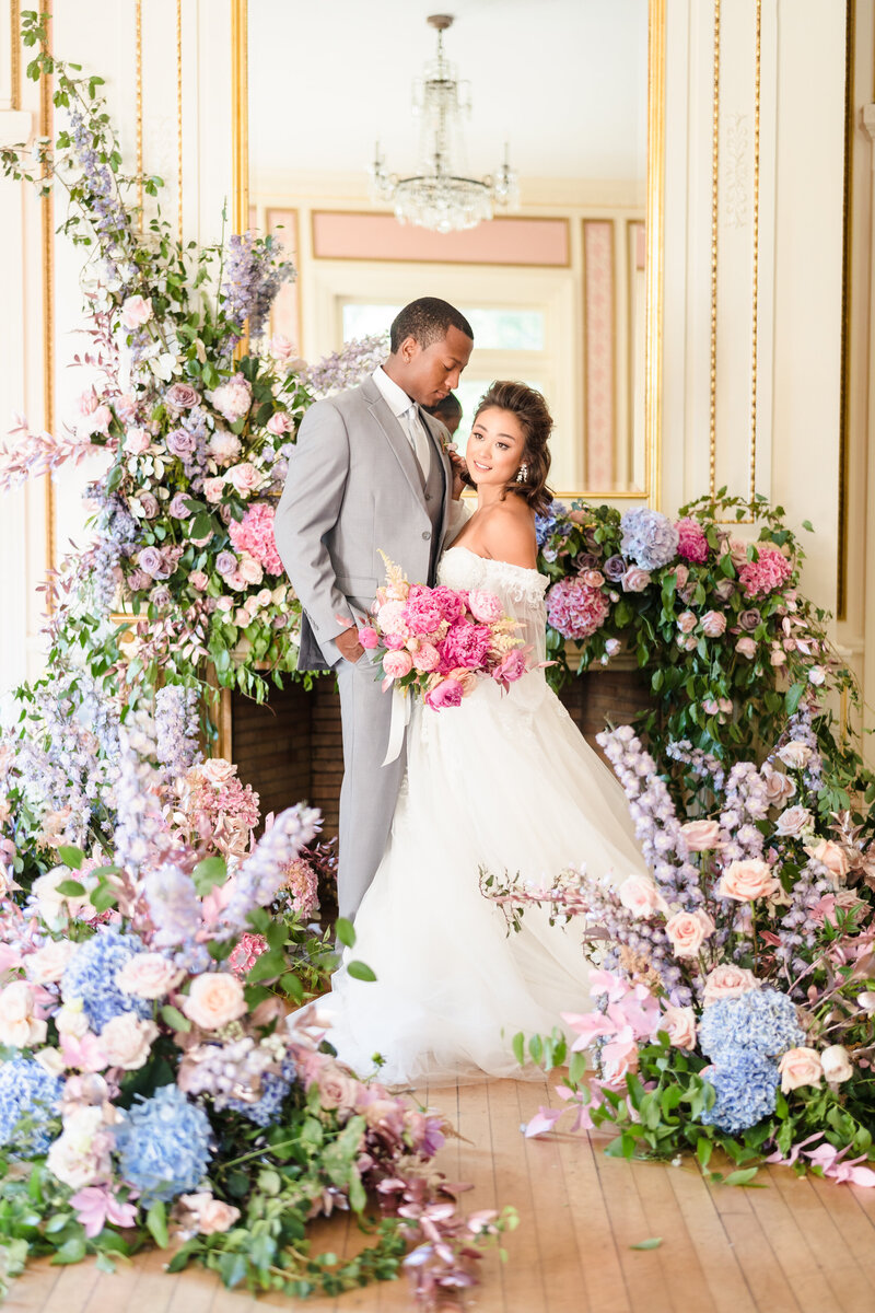 A bride and groom embrace in the middle of an elaborate array of pastel floral arrangements inside a historic ballroom