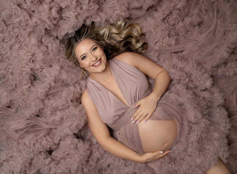 Pregnant Mom in a black bodysuit with an elegant black hat on. She is kneeling down, holding hat and showing her baby bump. Photo taken at Atlanta Maternity photo studio in Woodstock GA.