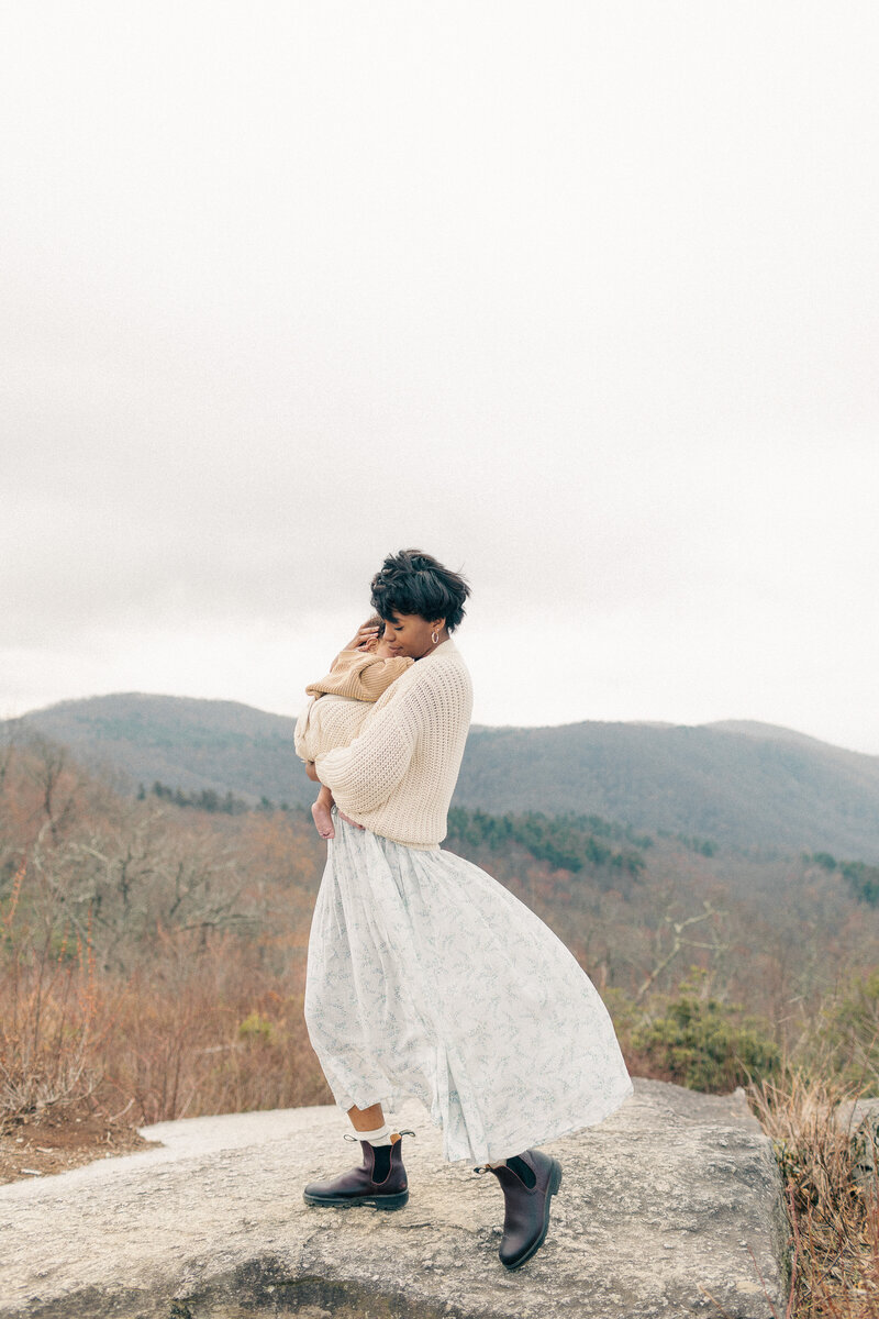 Mother gently holding her baby while standing on a rock ledge during an outdoor newborn session at a Kansas City overlook