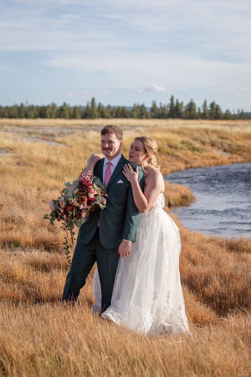 Bride in a lace wedding dress leans on the groom’s shoulder as they walk through a golden grass field beside a winding river, holding a bouquet of deep red and pink flowers.
