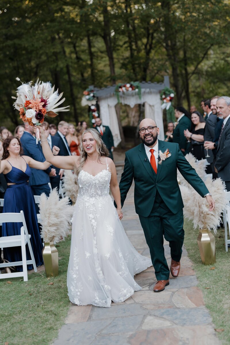 Bride and groom walking back up the aisle after their outdoor ceremony at Vecoma at the Yellow River near Stone Mountain, smiling and holding hands as guests celebrate around them.