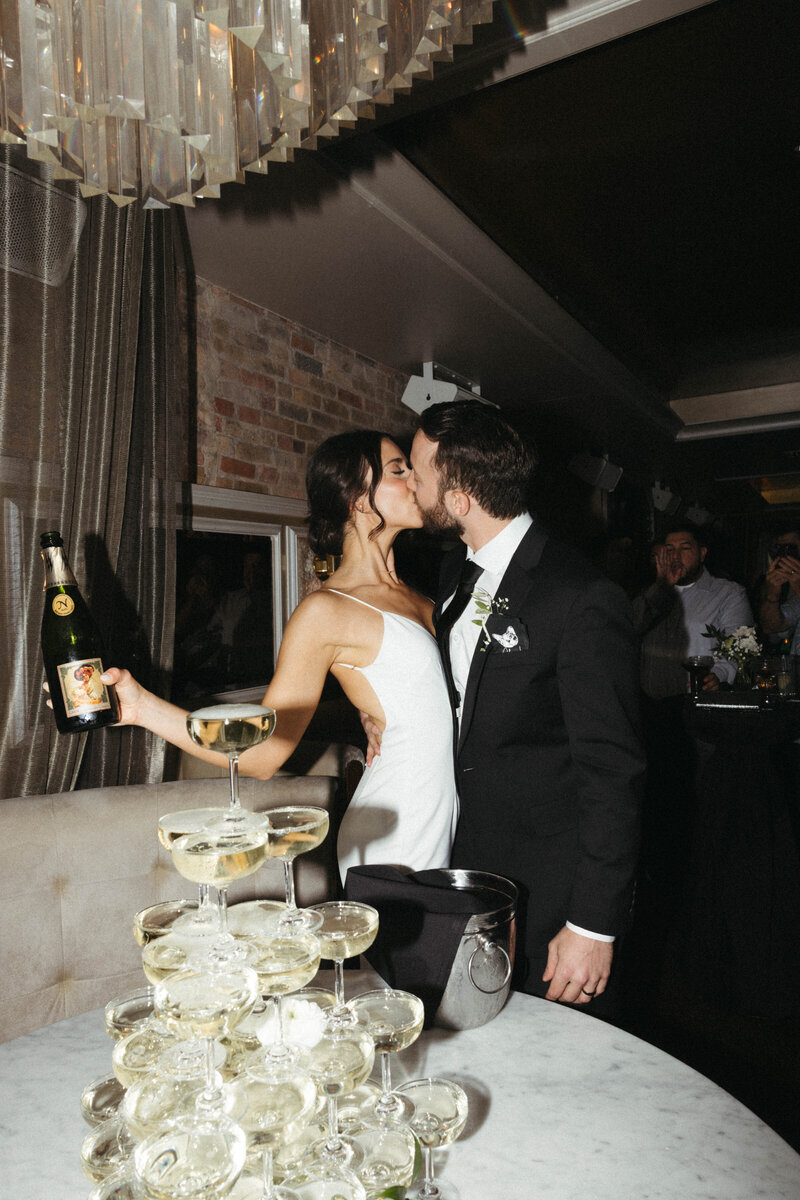 Newlyweds Kiss at Chicago wedding reception standing behind stacked, tiered glasses filled with champagne as bride holds the bottle.