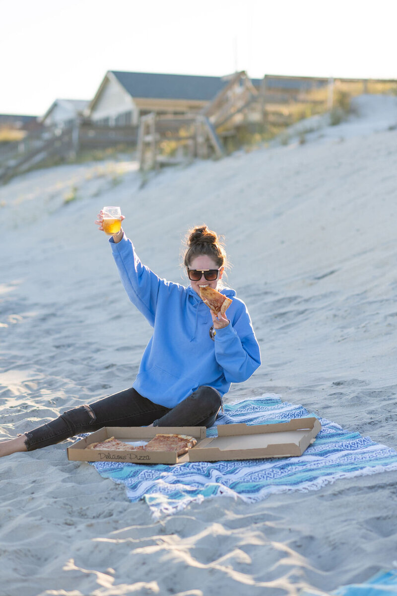 woman eats pizza and raises a glass while sitting in the sand