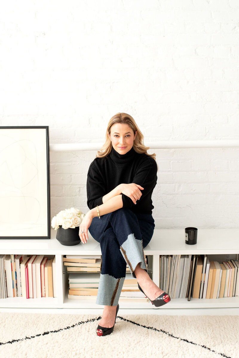 Laura Gatsos Young seated on a low bookshelf with legs crossed, smiling