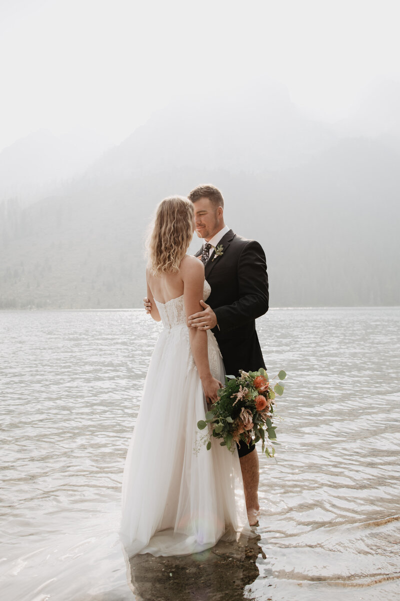 Grand Teton wedding pictures with bride and groom standing in the lake together while the sun shines over them captured by jackson hole photographers as the groom holds his brides hands