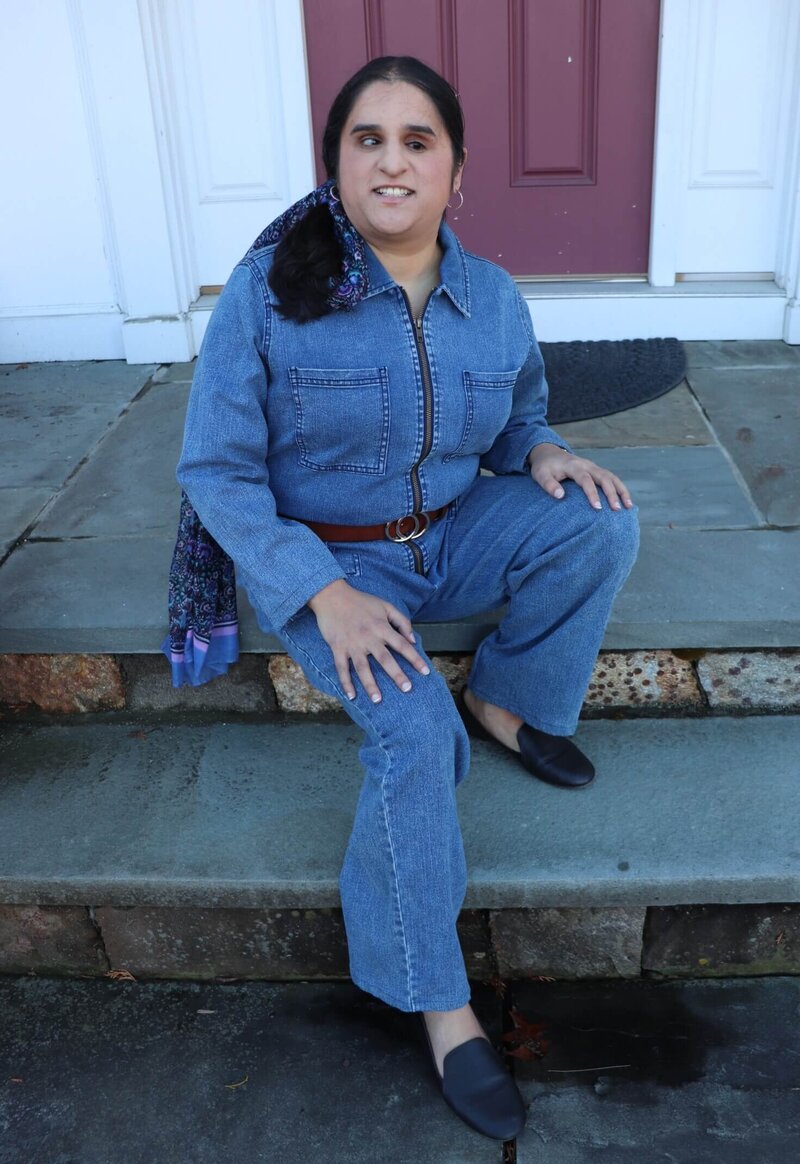 Ayesha of Writing Power-up and book coach for POC SFF writers smiling and sitting on steps in front of house with a light purple door