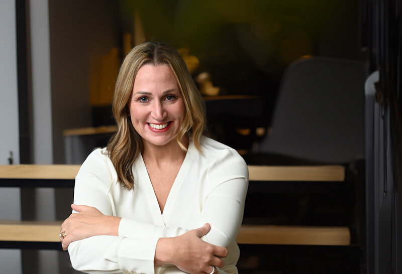 Jenn Cloutier professional headshot with bright smile, shoulder-length blonde hair, wearing cream-colored top against dark office background.