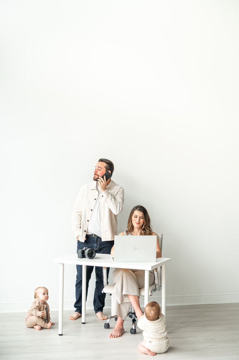 Kala is sitting at a white desk typing on her laptop while Reg is behind standing up talking on his phone. In front of them are their twin babies on the floor looking at them