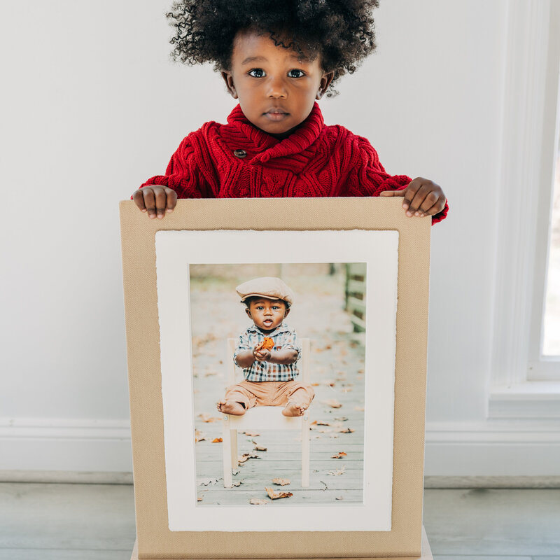 A toddler standing and holding a framed portrait of himself.