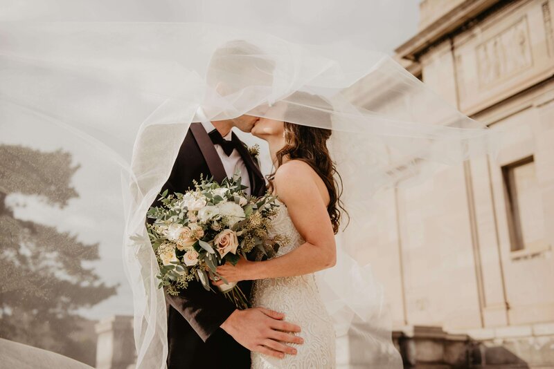 Bride and groom kissing under wedding veil while holding bridal bouquet of flowers