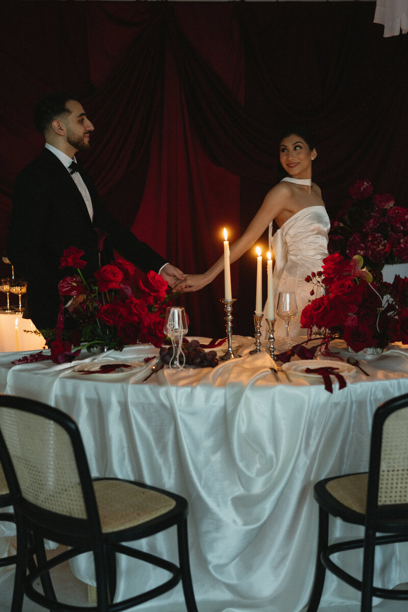 Couple beside a round table with white satin linen, red floral centerpieces, and taper candles in a moody reception setting.