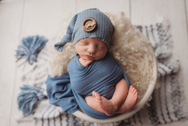 Baby newborn boy wearing a blue sleep cap swaddled in blue fabric with feet out and crossed and hand under chin