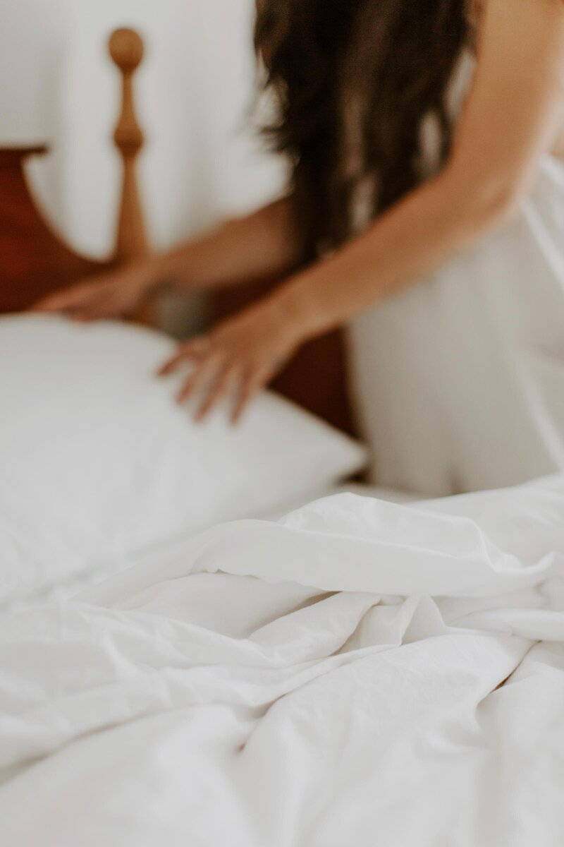 white bedsheets and pillow, wood dainty headboard, girl with long brown hair leaned over with hands on pillow