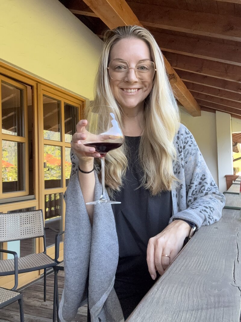 Marcie Muensterman is holding a glass of red wine. She has glasses on and long blonde hair. She is wearing a gray wrap over a dark top. She is leaning against a deck railing.