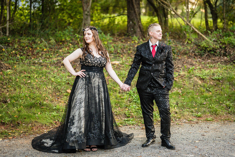 A queer couple hold hands and look away from each other, each of them have a stoic expression on their faces. The pair stand outside on a gravel road just a few steps away from their Roanoke Airbnb.