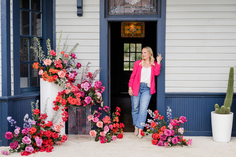 Bloom Bar founder in pink blazer posing in doorway with lush floral arrangements, reflecting confidence and creative direction in Austin floral design.