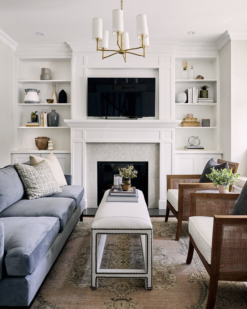 bright white timeless living room with built in floor to ceiling bookcases flanking a fireplace with a vintage rug and a soft blue velvet sofa with cane chairs designed by Lark & Linen interior design