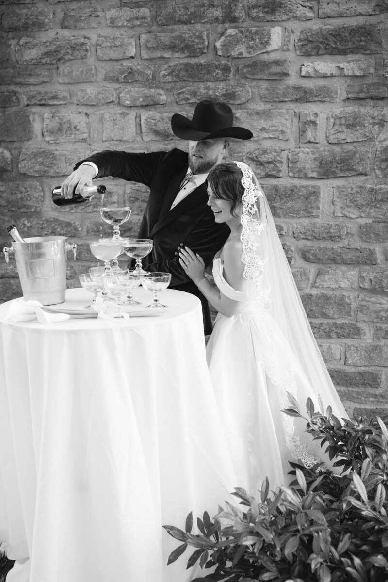 Black and white image of bride and groom in cowboy hat pouring champagne at Nashville wedding